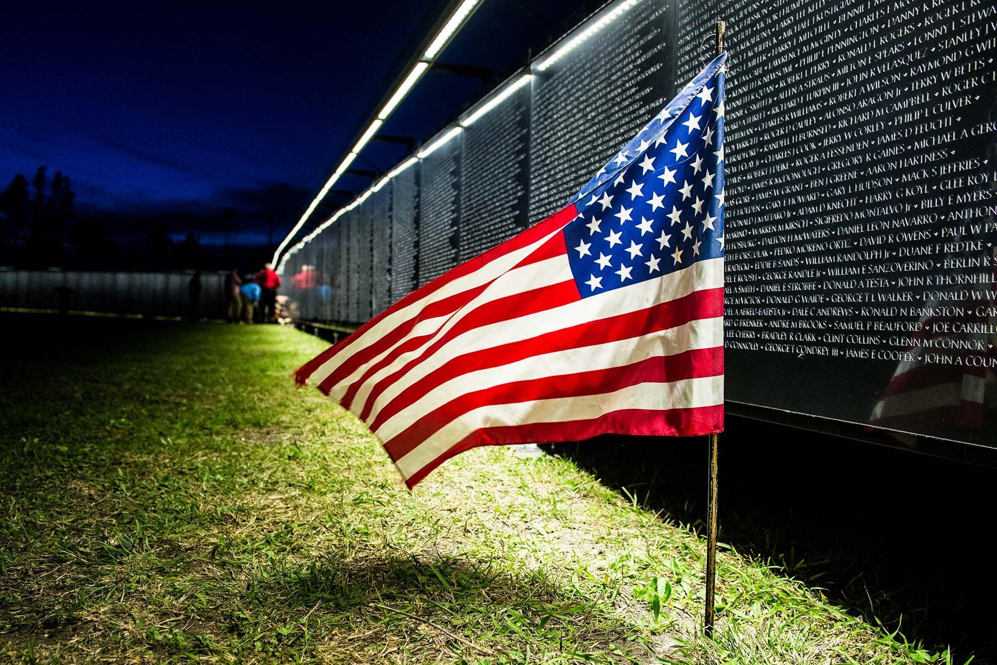 An American flag stands before an illuminated panel of The Wall That Heals at night
