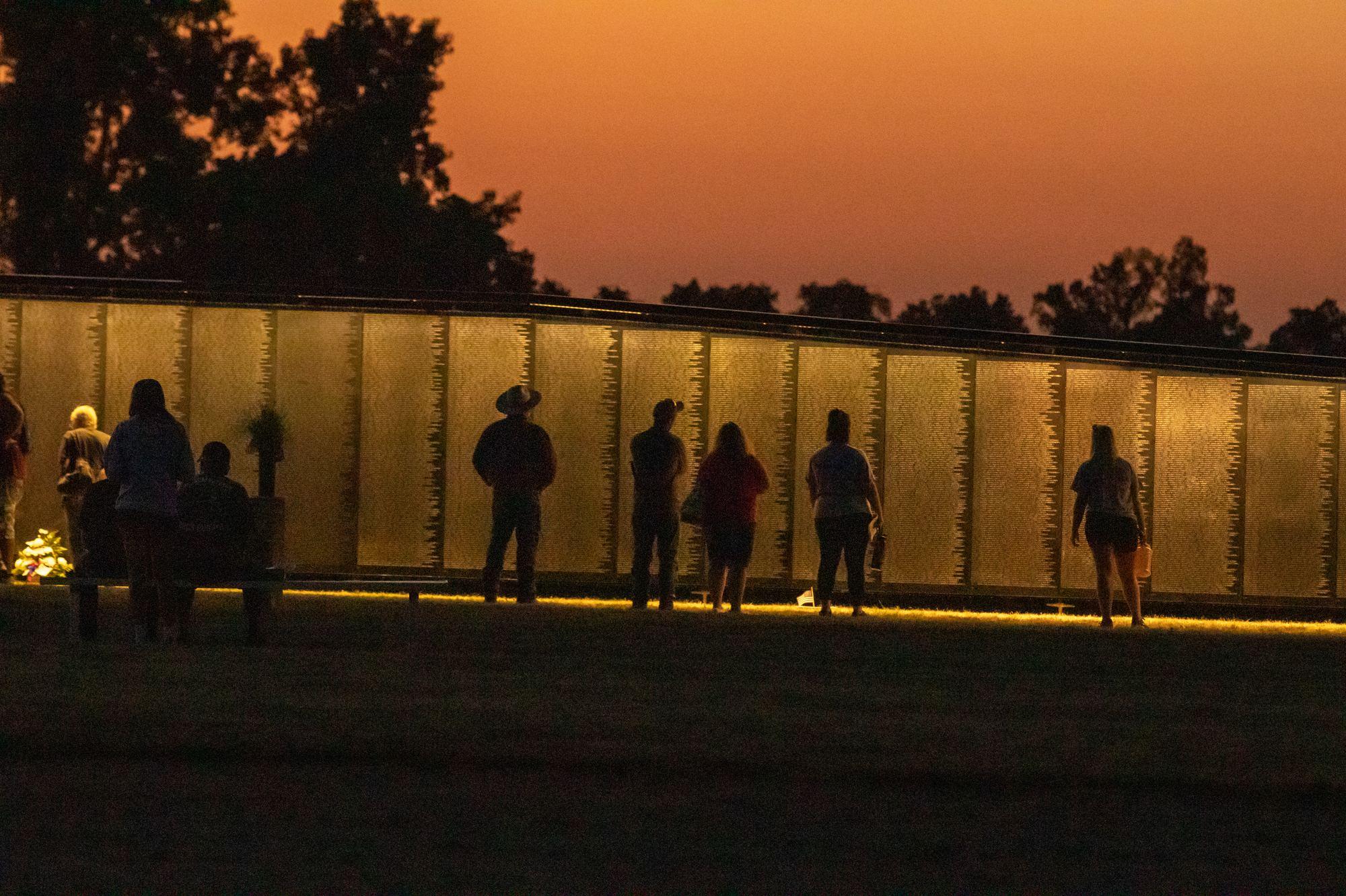 Visitors silhouetted against The Wall That Heals glowing amber at night