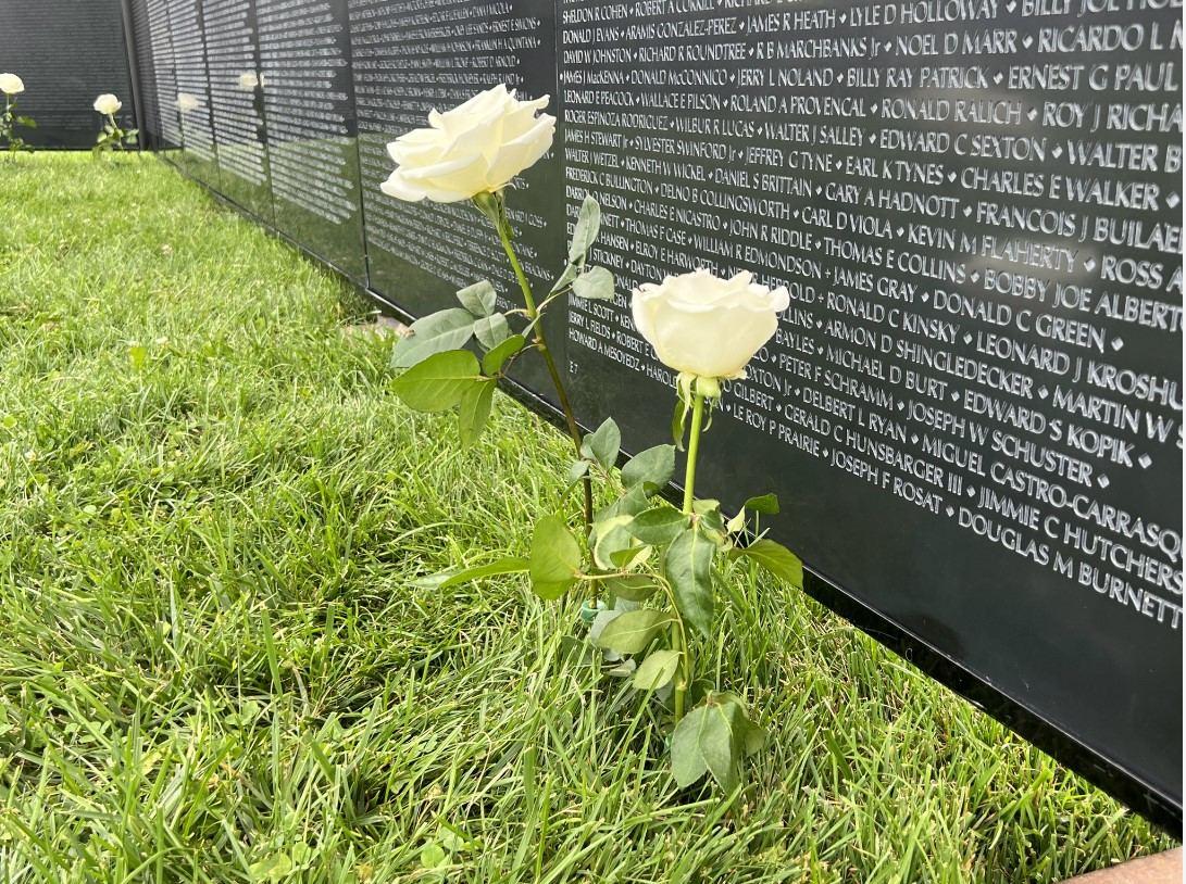 Two white roses lean against the names engraved on The Wall
