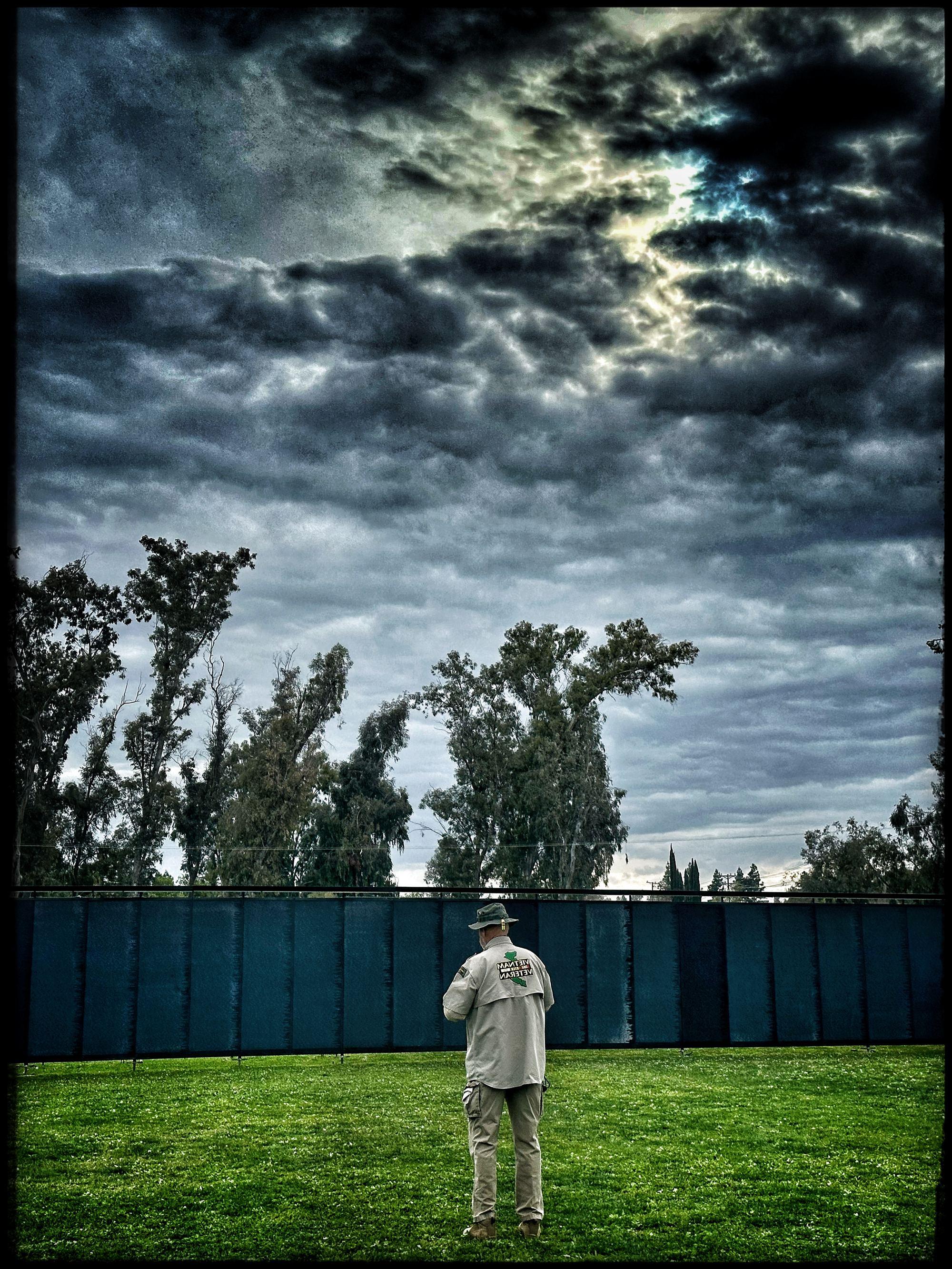 A Vietnam veteran stands before The Wall That Heals at dusk beneath a stormy sky